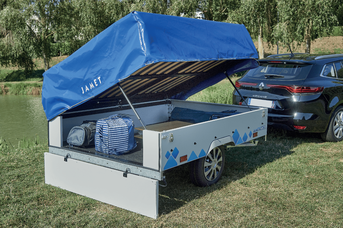 A Jamet trailer parked on grass beside a black car, with a blue canopy cover labeled "JAMET", containing bags and equipment, near a river and trees.