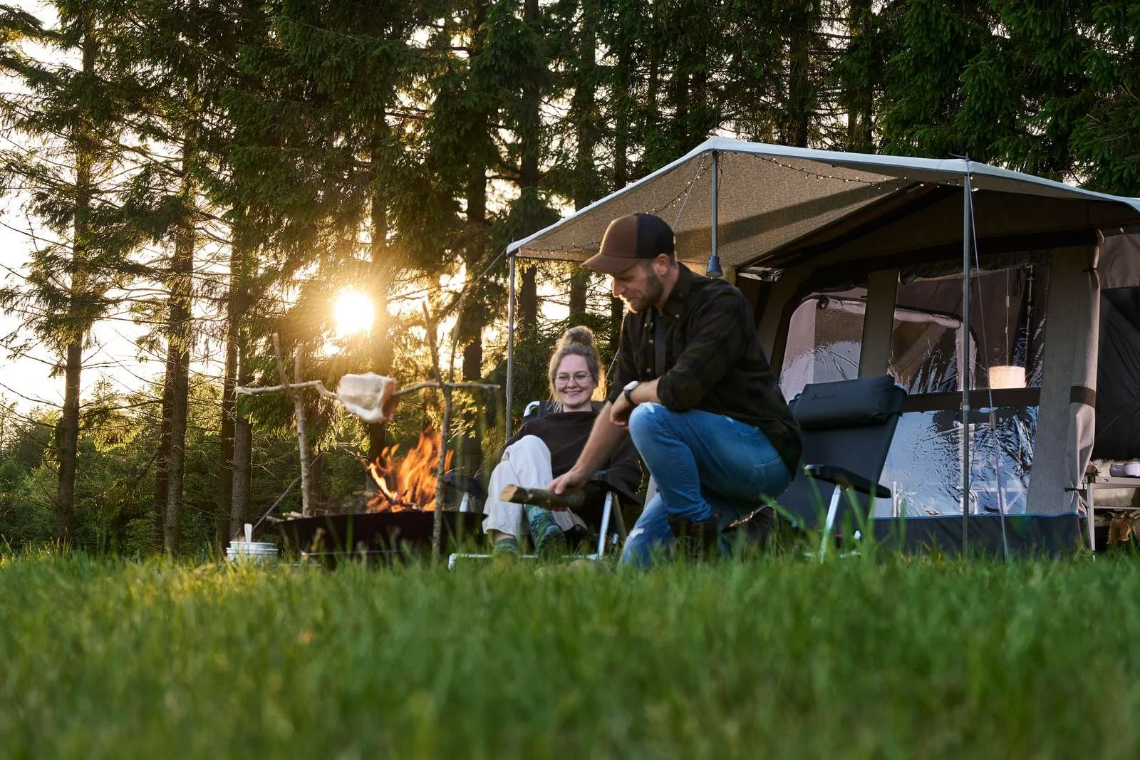 Two people camping outdoors near a Camp-let Earth trailer tent, with one person grilling marshmallows over a campfire and the other sitting nearby smiling as the sun sets behind trees.