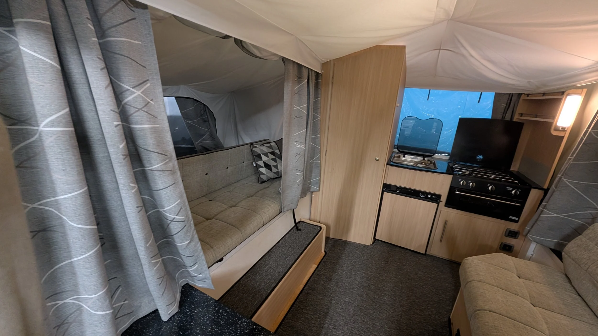 Interior of a compact camper or RV with a small kitchen, a sofa, a window with curtains, and wood cabinetry.