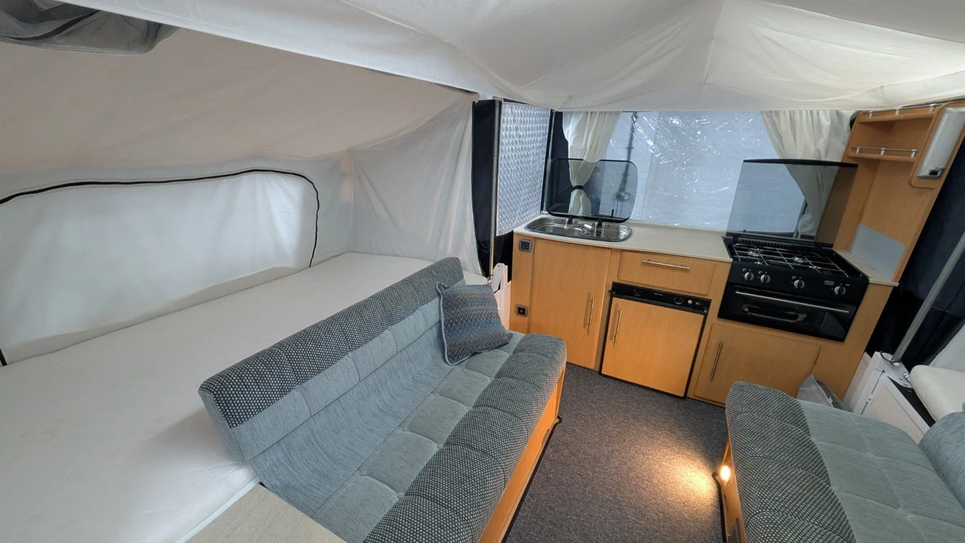 Interior of a small camper or RV showing a bed, kitchen area with sink, stove, and cabinets, and a small sofa.
