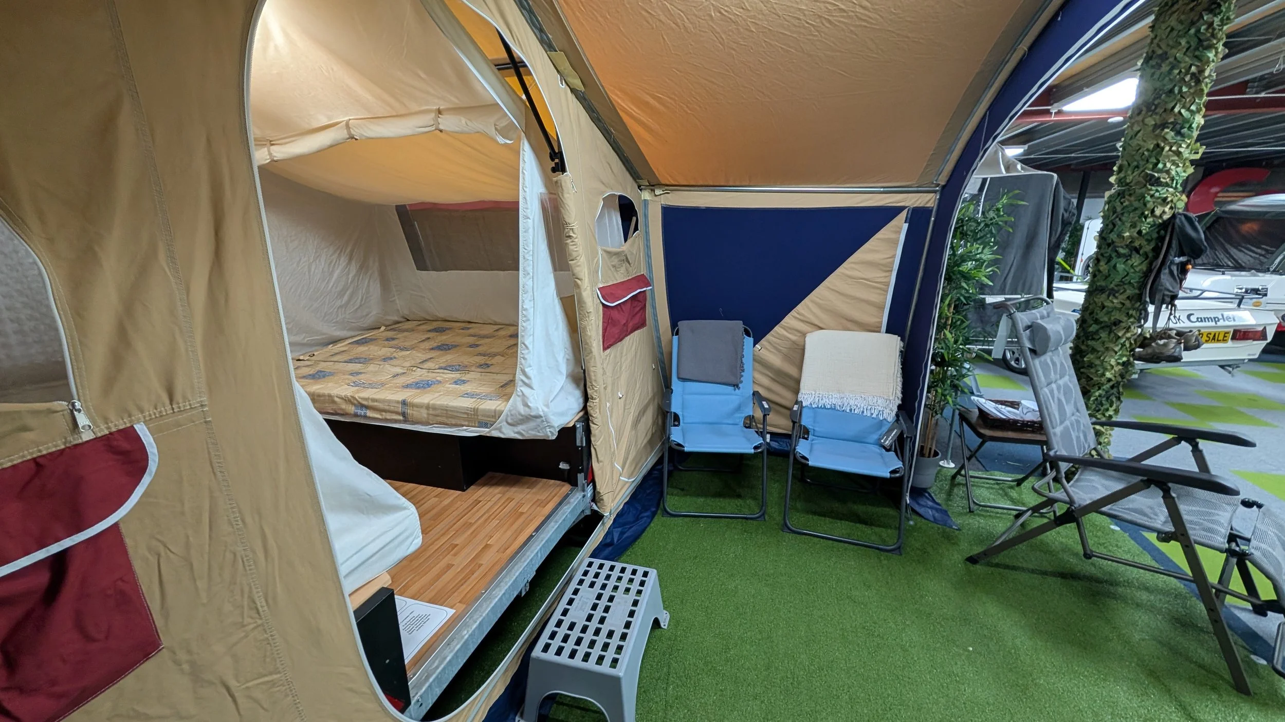Display of a camping tent with outdoor furniture including chairs and plants at an indoor showroom.