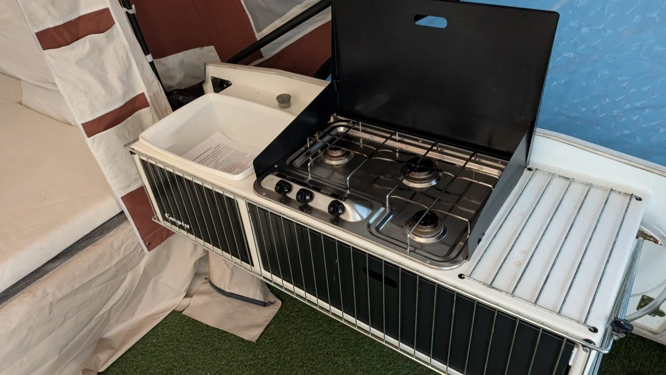 A vintage camping stove with three burners, black lid, and handles, situated next to a small sink on a camping or RV setup, with bedding and foam mat visible in the background.