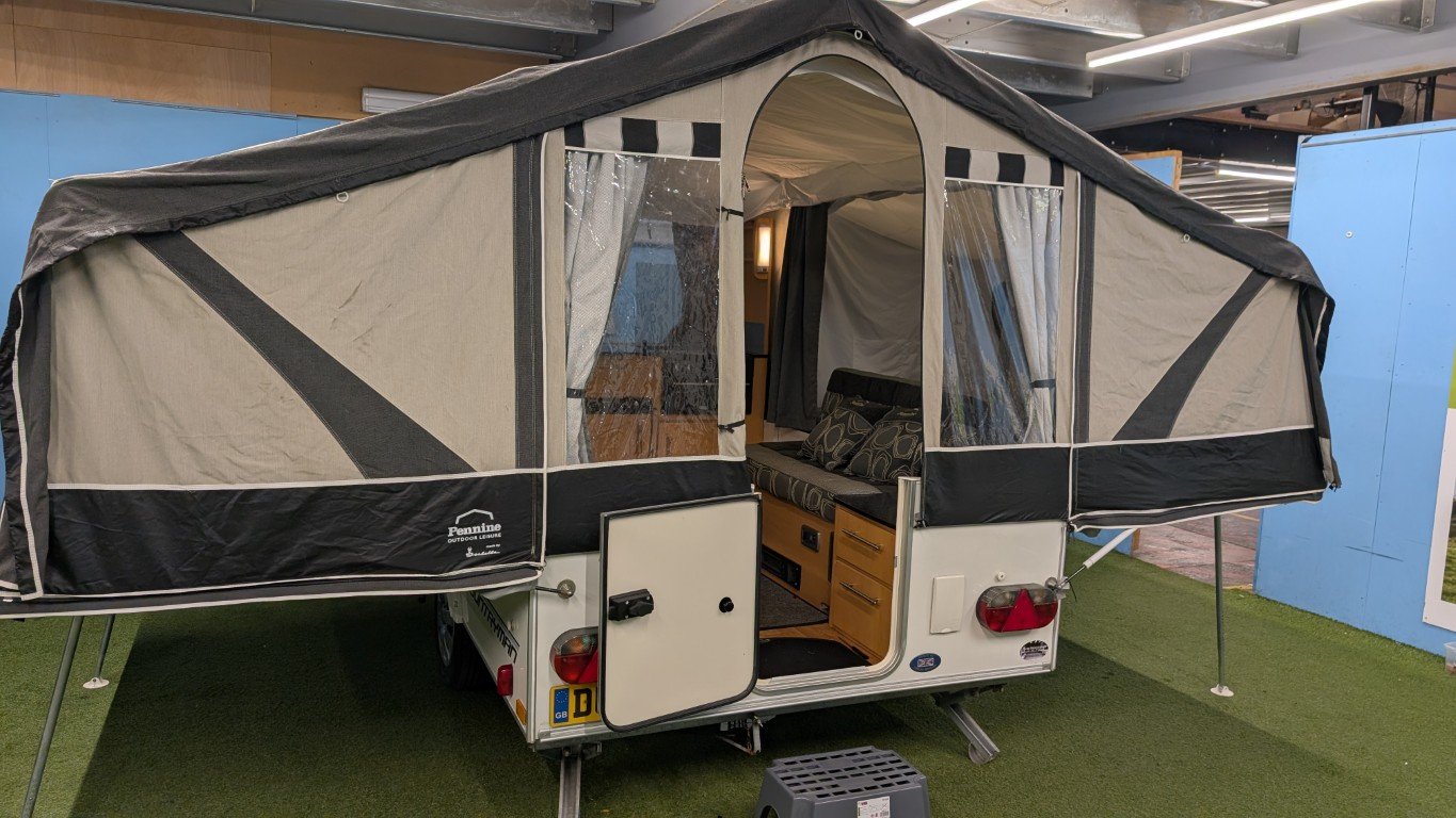 A compact camping trailer with an extended tent area, open door revealing an interior with a bench and curtains, set on artificial grass inside a building.