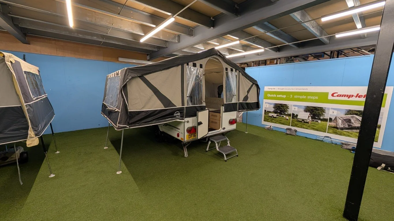 An outdoor camping trailer with a pop-up tent extension, set up indoors with artificial grass flooring and a display board showing quick setup steps.
