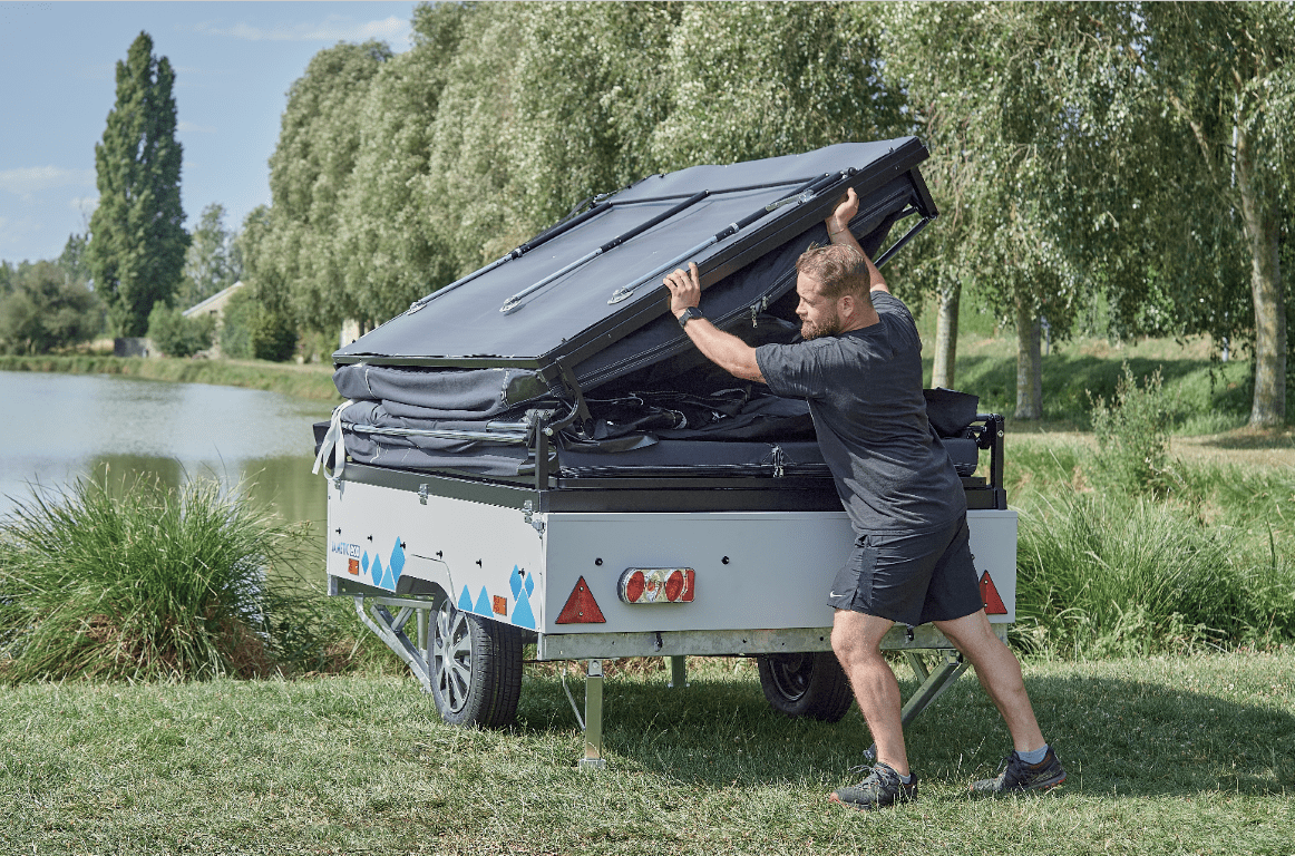 Man setting up a Jamet camping tent trailer near a lake surrounded by trees.