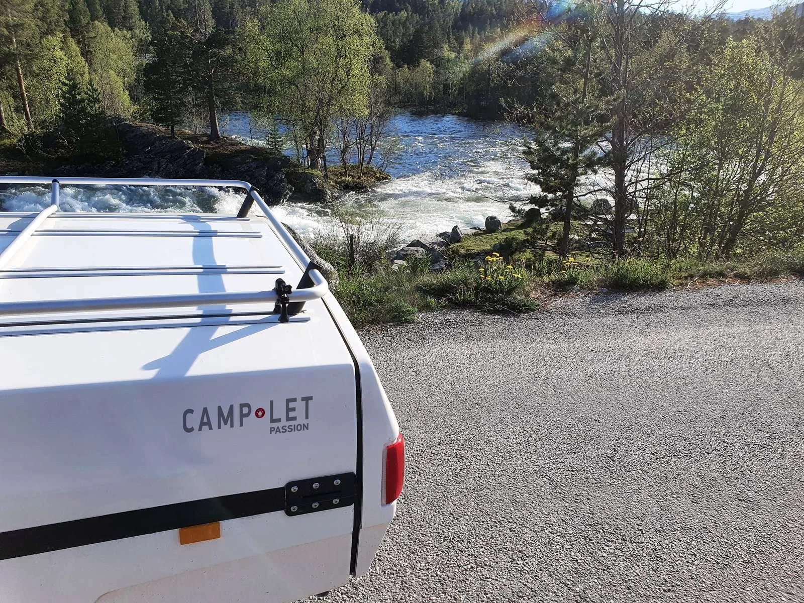 The back of a white Camp-let trailer Tentparked on a gravel road near a river with trees and rocks, a rainbow, and a bright sky in the background.