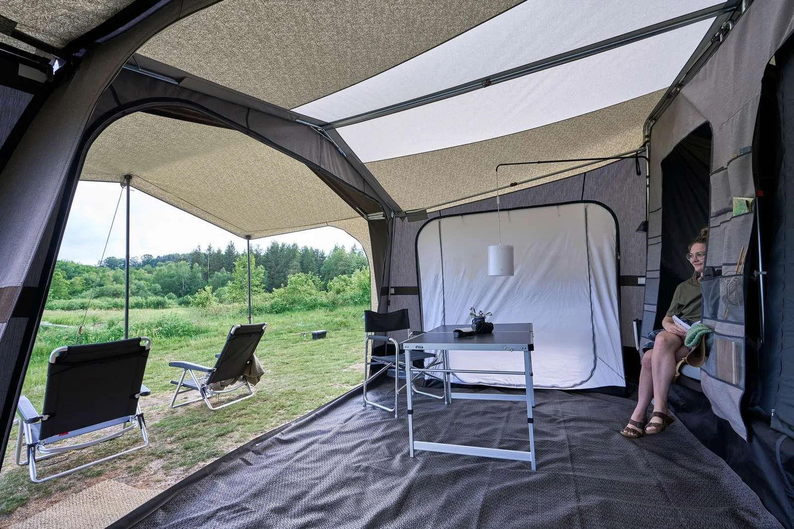 View from inside a camping tent with a view of green grass and trees outside. The tent has a gray and white interior, a small table with a plant, a woman sitting on the right side, and outdoor chairs facing the scenery.