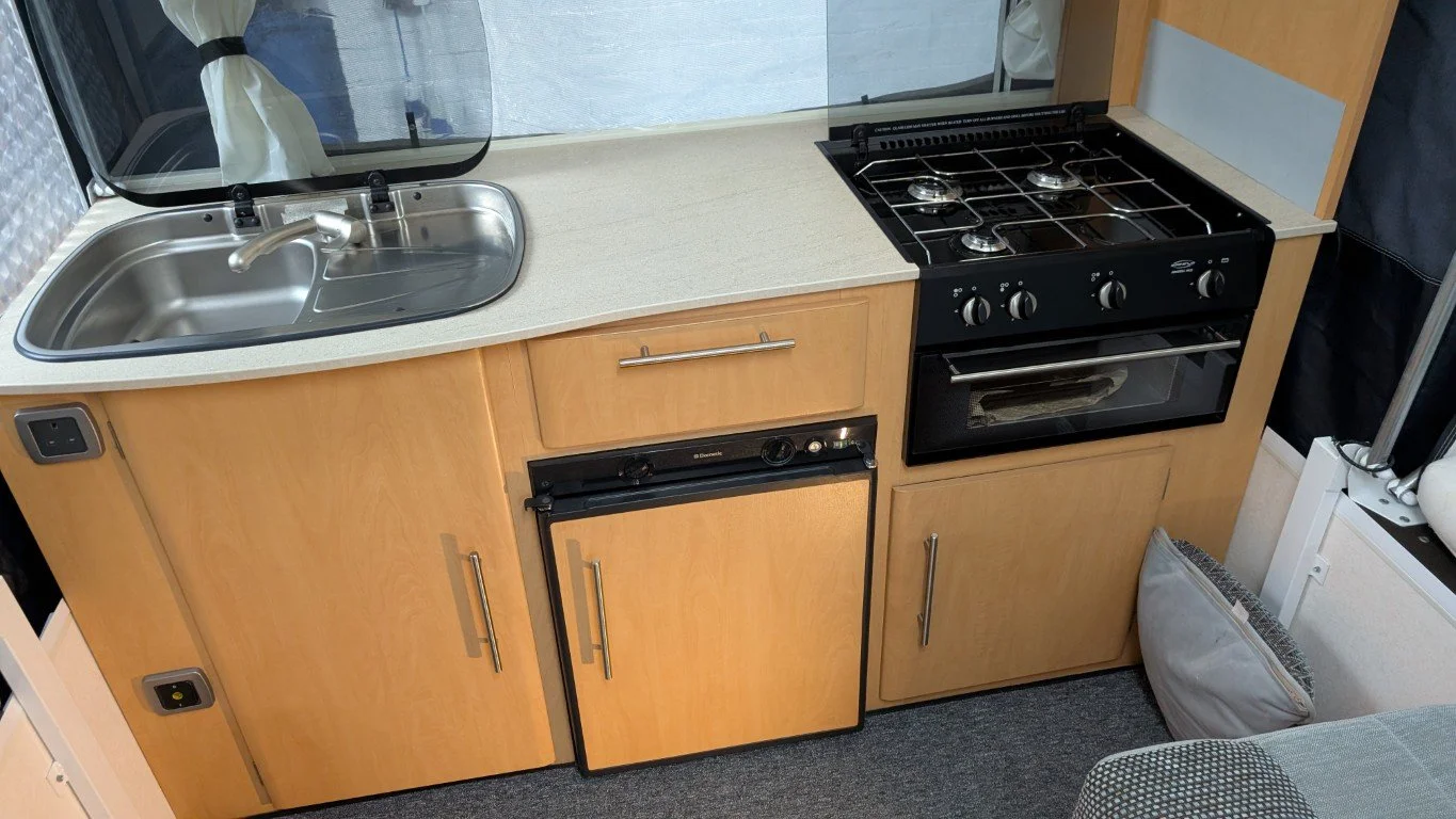 Small kitchenette with a stainless steel sink, a compact refrigerator, a four-burner gas stove, and wooden cabinets, with a curtain, a window, and a pillow visible.