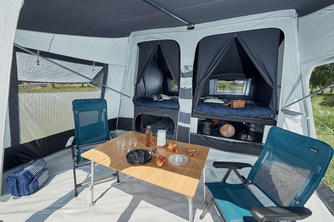 Interior of a Jamet camping tent with two screened sleeping areas with beds, a small wooden table with glasses, bottles, and food, and two blue camping chairs.