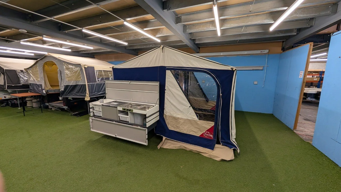 Display of various camping tents and outdoor gear inside a store, with artificial grass flooring and blue wall panels.