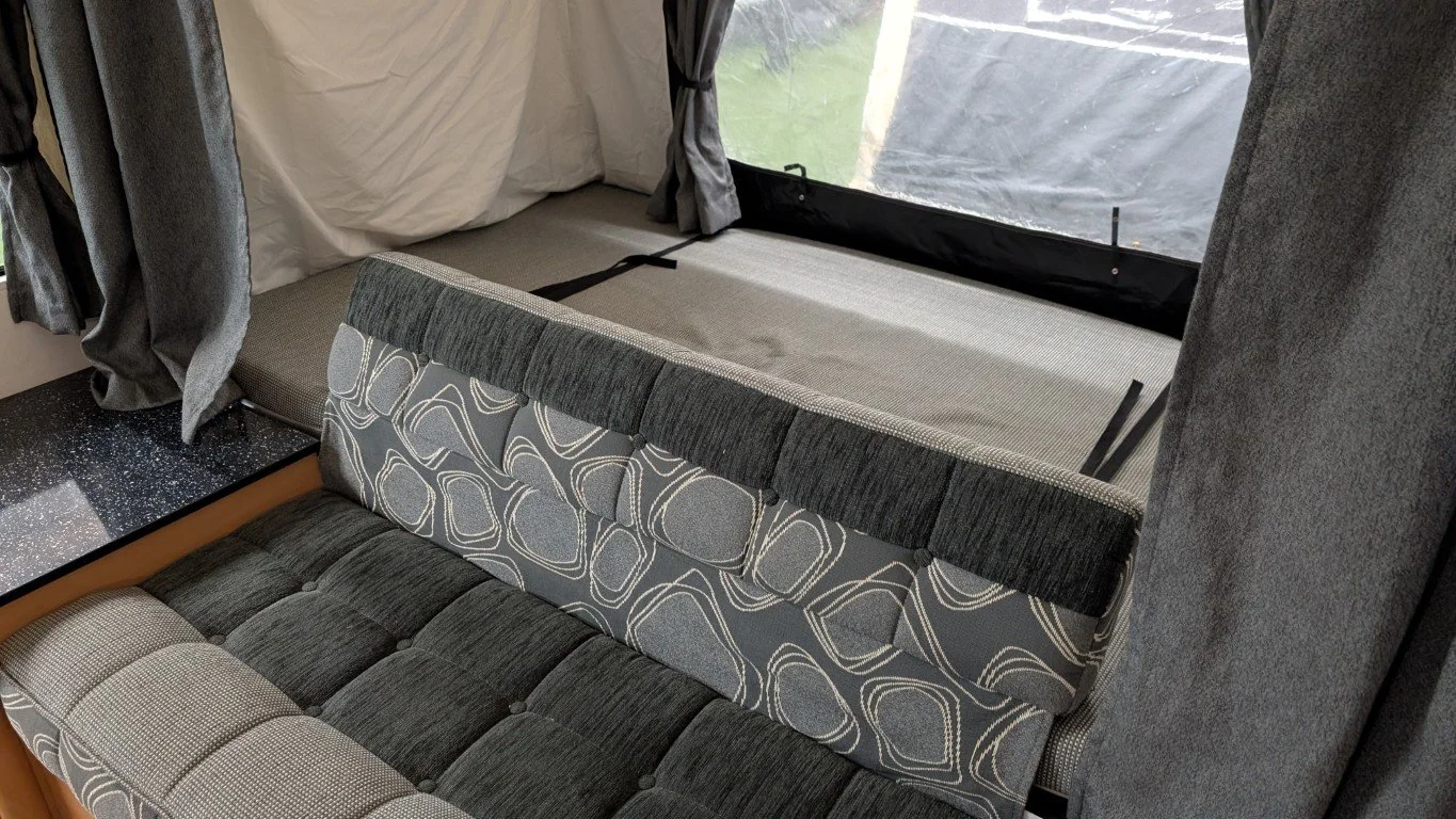 Inside of a camper contains a fold-out sofa with patterned cushions and a raised sleeping area with a window and curtains.
