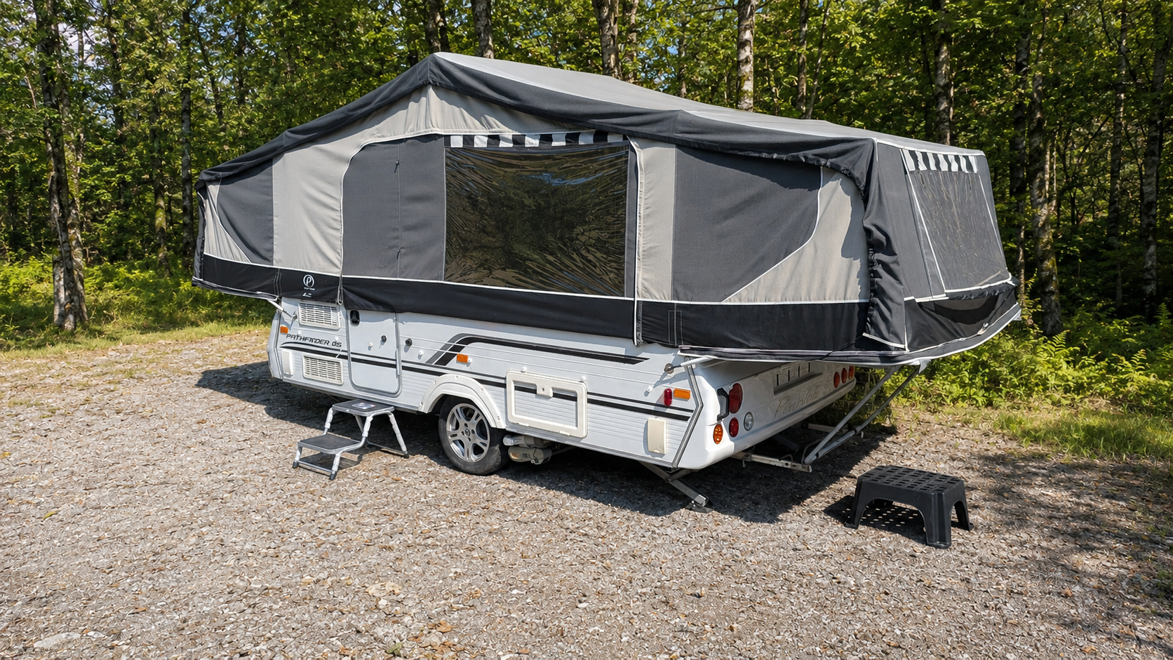 A travel trailer camper with an expanded tent cover, parked on a gravel lot next to a wooded area. A small step ladder is placed at the door.
