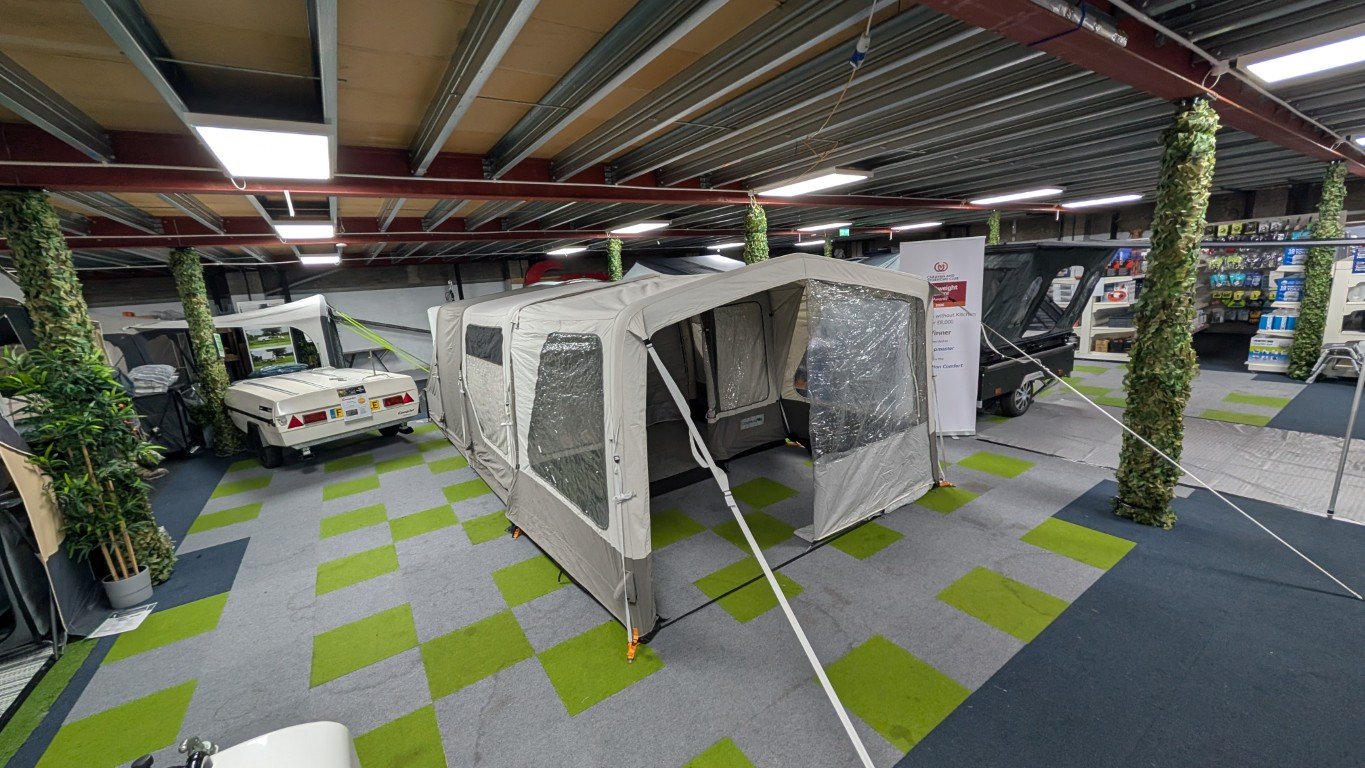 Indoor showroom displaying camping and outdoor gear, including a large beige tent with transparent windows, surrounded by miniature vehicle models and decorated with hanging greenery.