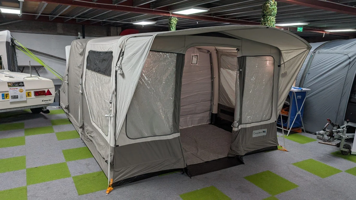 A grey and white camping tent with a large open entrance, set up indoors on green and grey checkered flooring, next to a white camper trailer in a showroom or display area.