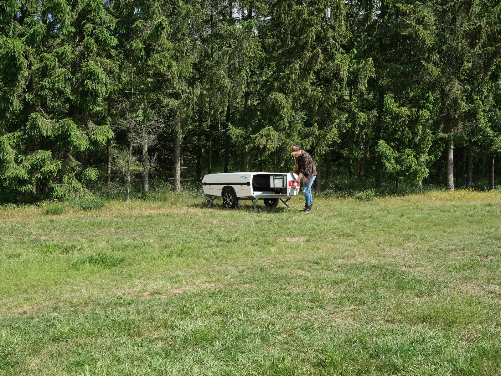 A person preparing a Camp-let  trailer in a grassy field with trees in the background.