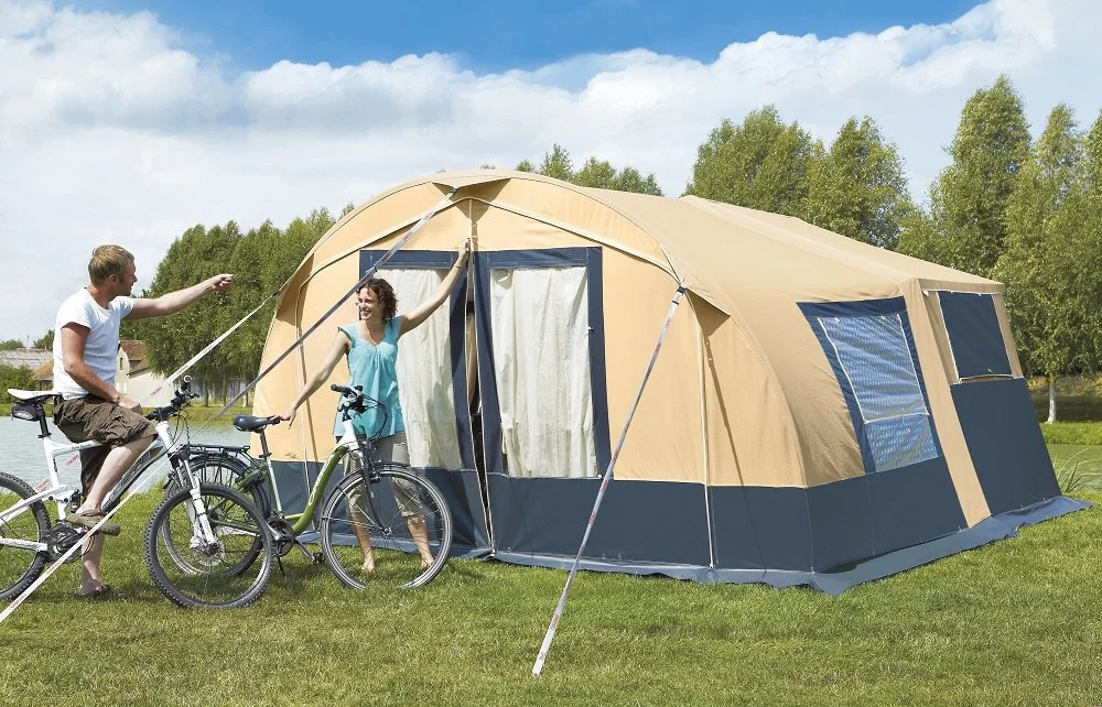 A man and a woman are standing outside a large beige and blue Camplair tent near a lake, with bicycles nearby. The man is on a bicycle, pointing, while the woman is standing at the tent entrance smiling.