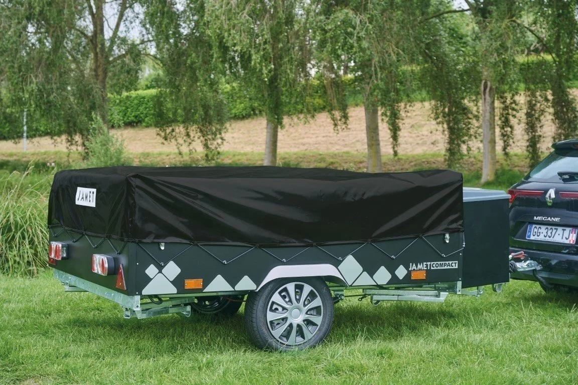 A black Jamet Compact trailer with a cover, parked on grass near a tree and a black car.