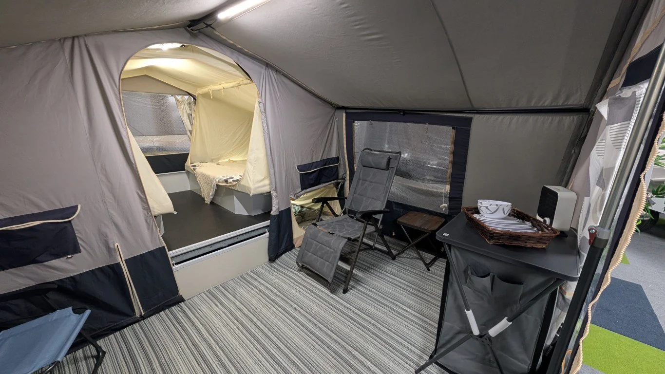 Inside a camping tent with a striped floor, a foldable chair, a small table, a basket with plates and bowls, and a window on the side. The tent has a screened opening revealing a sleeping area with a bed covered in white sheets.