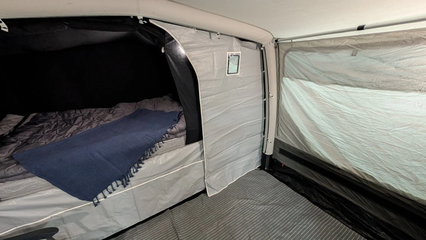 Interior of a camping tent with a sleeping area visible, featuring a bed with gray bedding and a blue throw blanket. The tent walls are light-colored fabric.