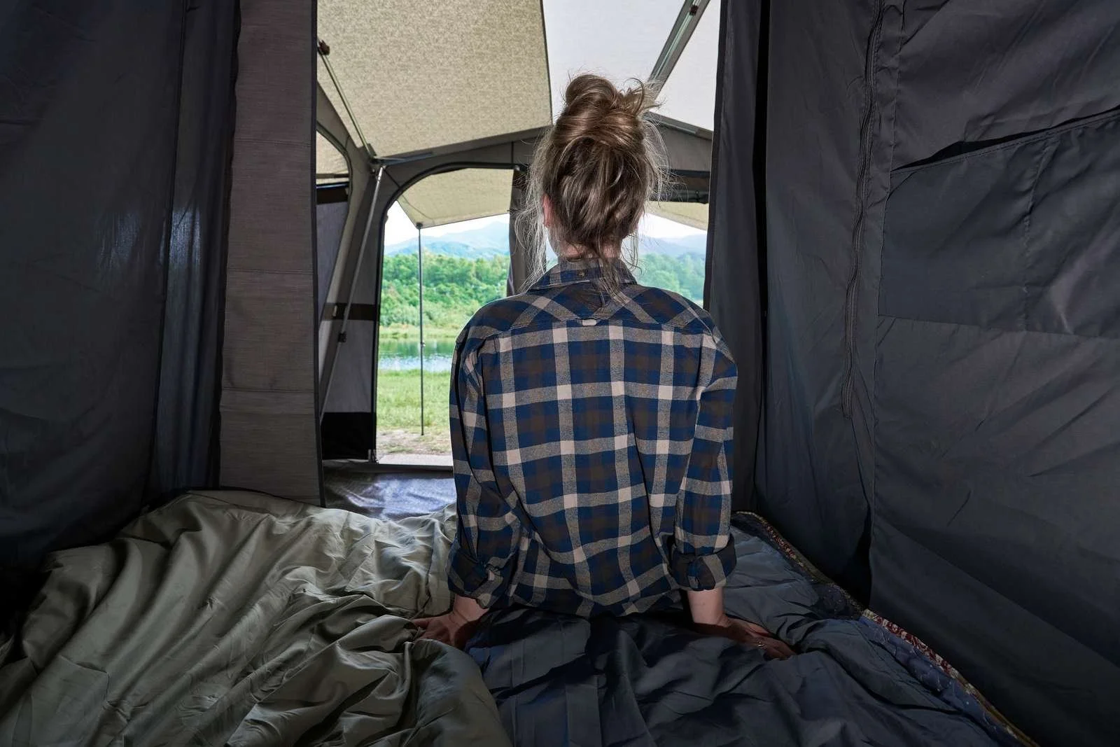 Person sitting inside a camping tent, facing outside, overlooking a lake and green scenery.