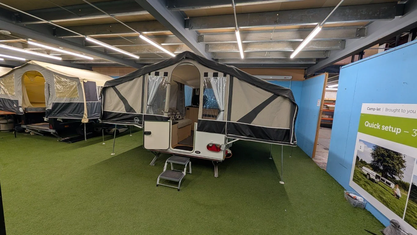 A pop-up camper trailer displayed indoors on green artificial grass with a small set of steps leading inside. There is another tent-style camper trailer adjacent on the left and informational signage to the right.