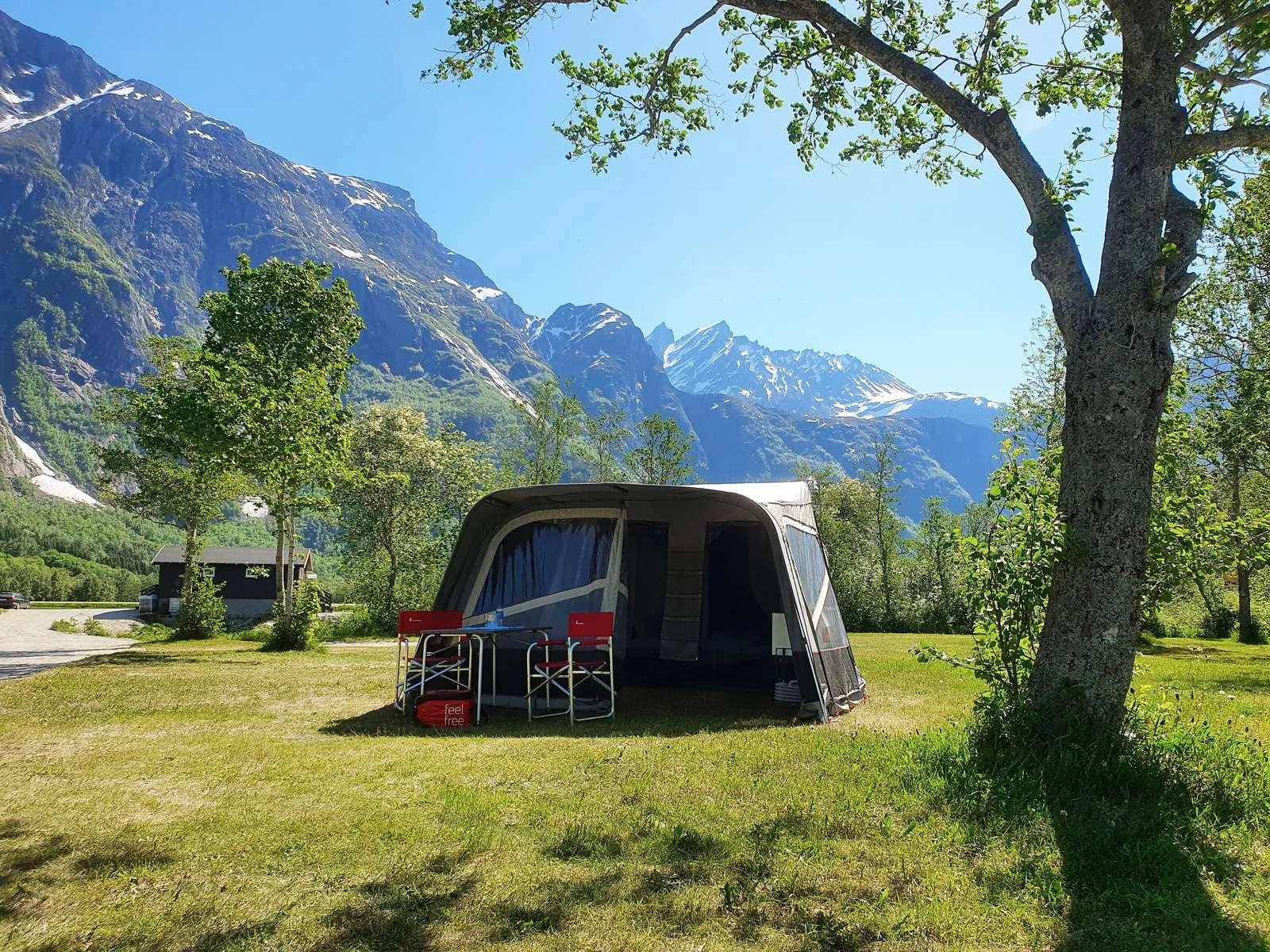A camping scene with a Camp-let set up on a grassy area surrounded by trees, with mountains in the background and clear blue sky overhead.