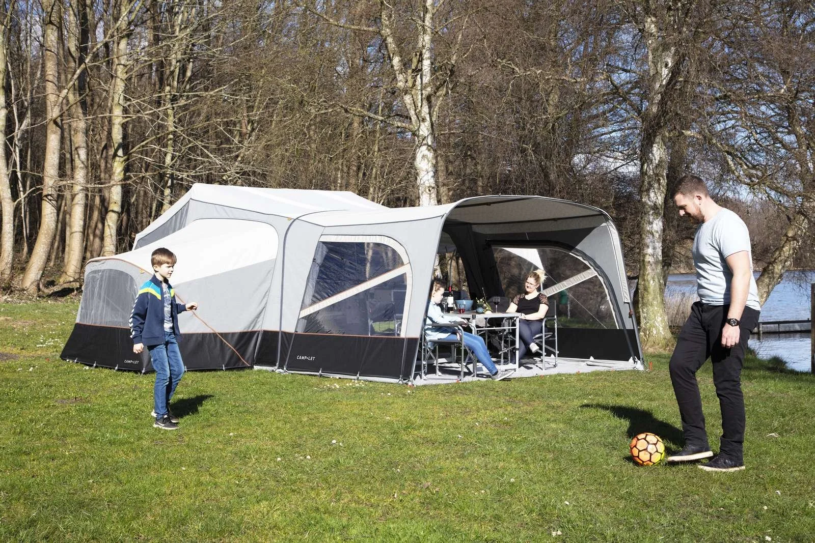 Family enjoying outdoor activities near a Camp-let tent by a lake with trees in the background. One person is playing with a soccer ball, while others are sitting at a table inside the tent.