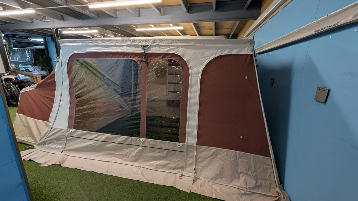 A beige and brown portable camping tent set up indoors on a green carpet, with clear plastic windows and a zippered door.