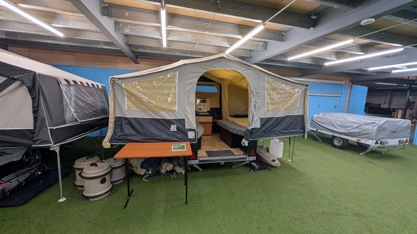 Display of various camping equipment including a tent, a trailer, and camping gear in an indoor showroom with artificial green grass flooring.