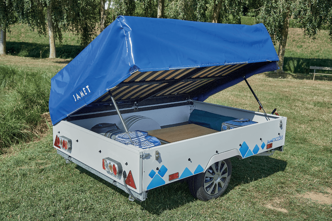 Jamet trailer with a blue canopy, parked on grass near trees. The trailer contains some luggage and a wooden surface inside.