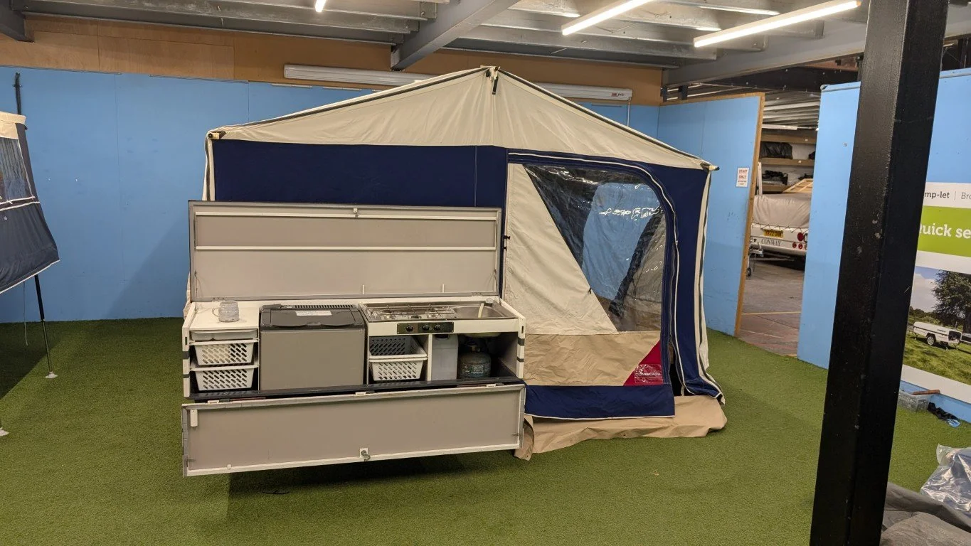 Indoor display of a camping tent attached to a kitchenette with cabinets and a gas tank, set on artificial grass with promotional banners in the background.