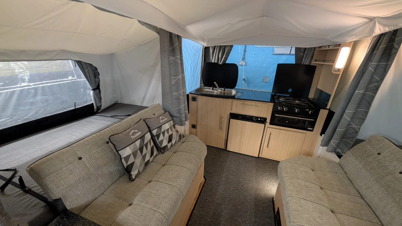 Interior of a camper or small living space with two light-colored sofas, a small kitchen area with a sink, mini fridge, and stove, and a window with curtains.