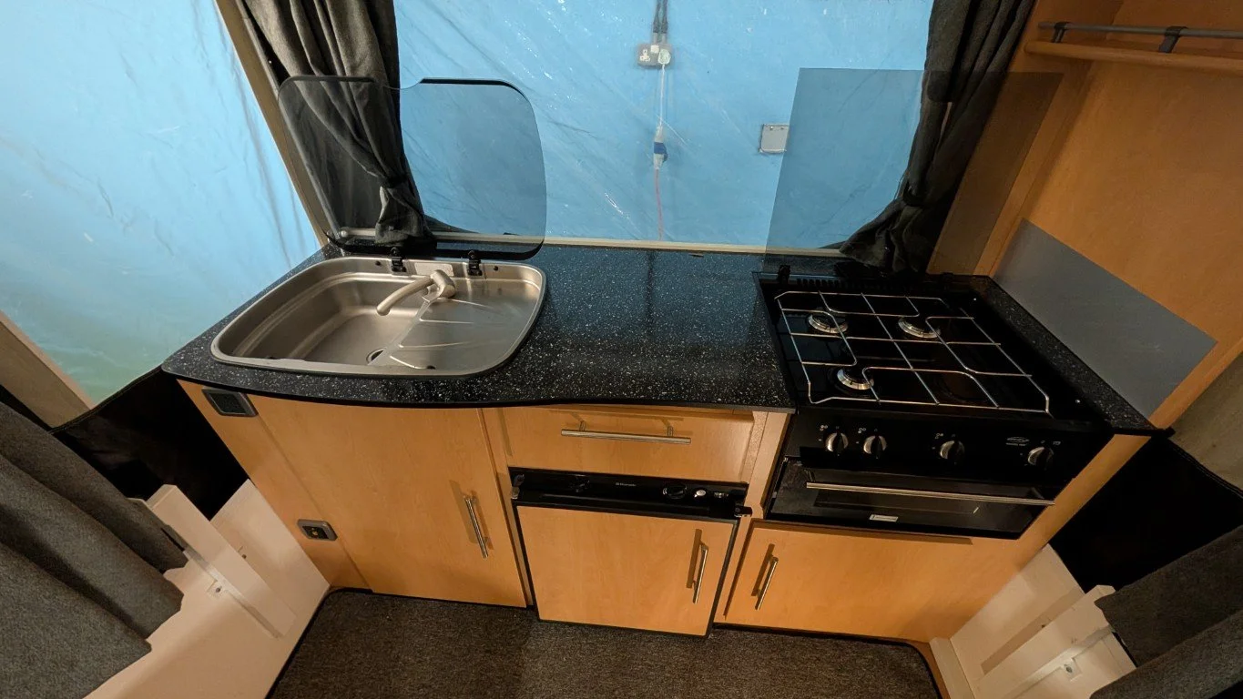 Compact kitchen with a black speckled countertop, a stainless steel sink with a glass cover, a black gas stove with four burners, wooden cabinets, a window with dark curtains, and a small refrigerator below the counter.