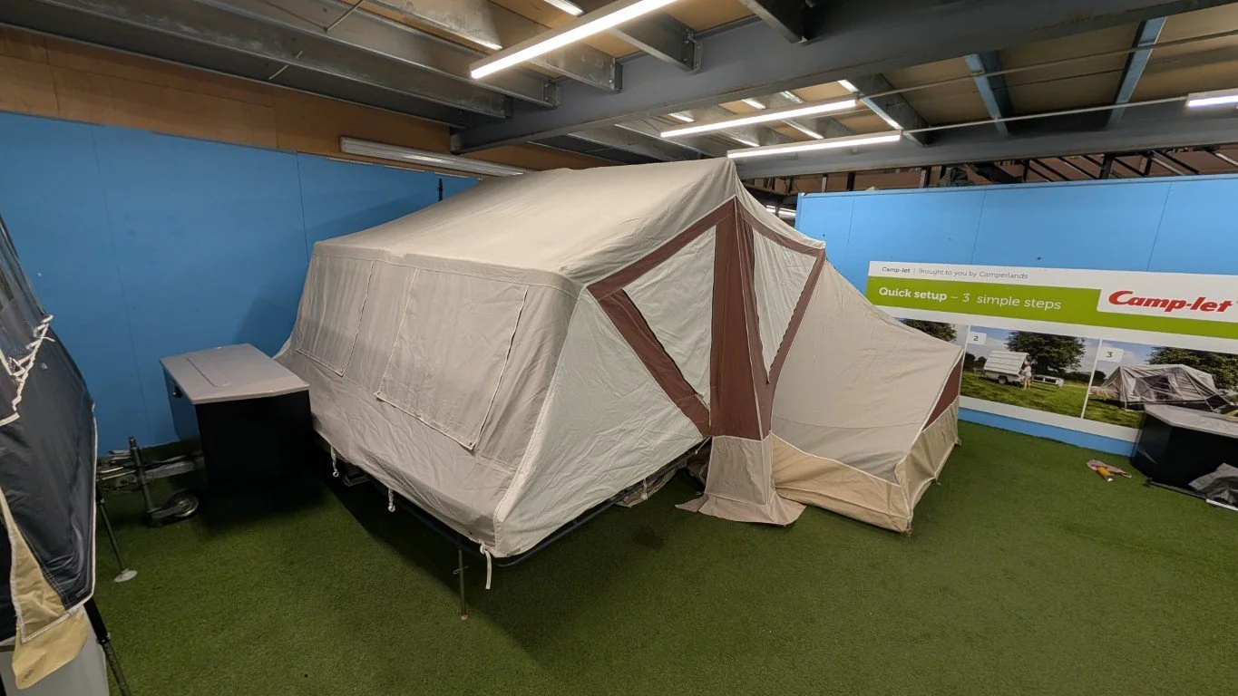 A large beige and brown camping tent set up indoors on artificial grass, with a blue wall and a sign in the background.