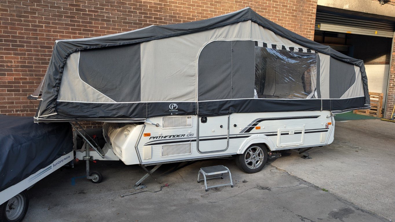 A pop-up camper trailer titled 'Pathfinder Q5' is parked on a concrete surface next to a brick wall, with a lowered roof and an enclosed canopy.