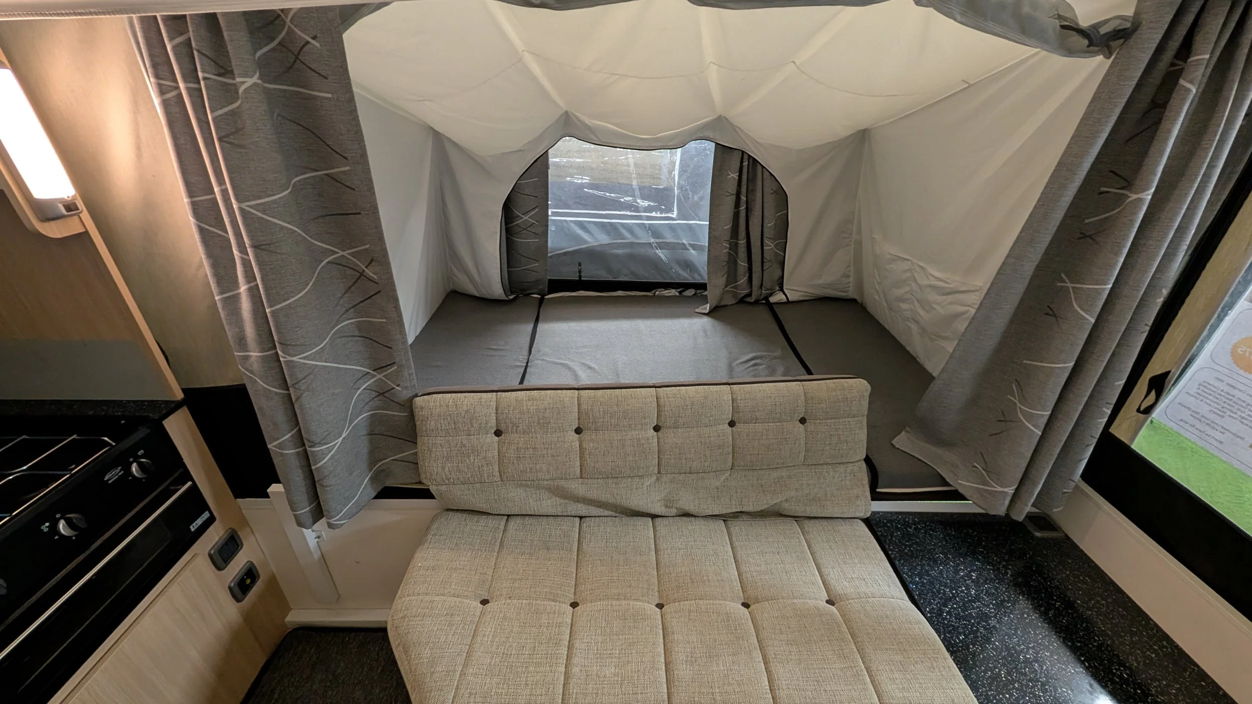 Interior view of a camper or small RV with a bed area, window with curtains, and a beige cushioned bench or sofa in the foreground.