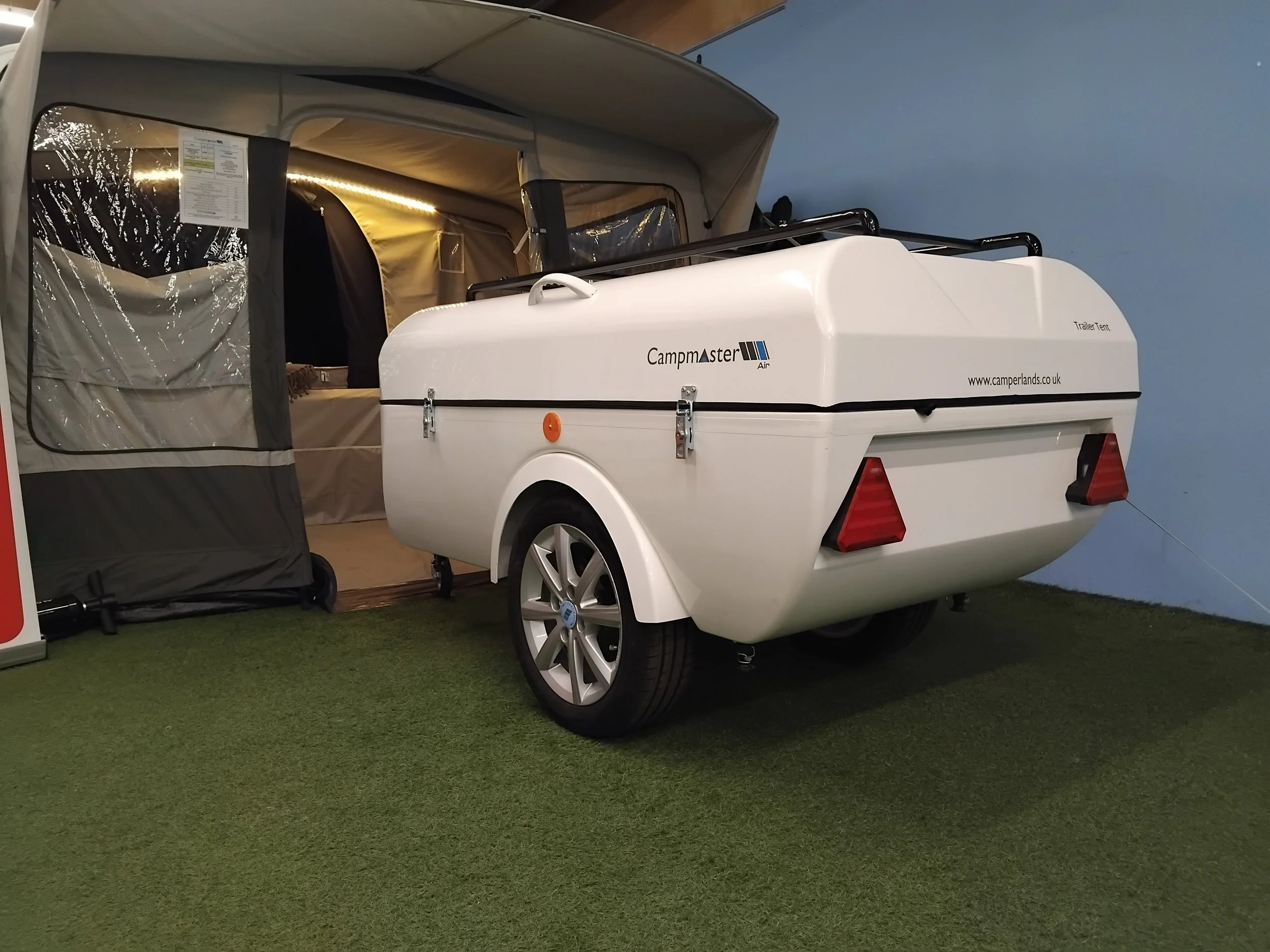 White camper trailer connected to a tent setup indoors on artificial grass, with a blue wall in the background.