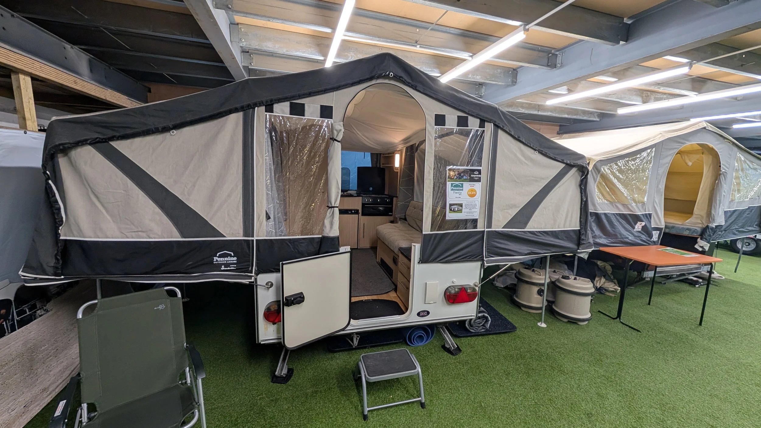 A compact travel trailer with a pop-up tent set up inside a store, featuring a kitchenette, a small couch, and a dining table. The trailer is displayed on artificial grass with some camping chairs and accessories around it.