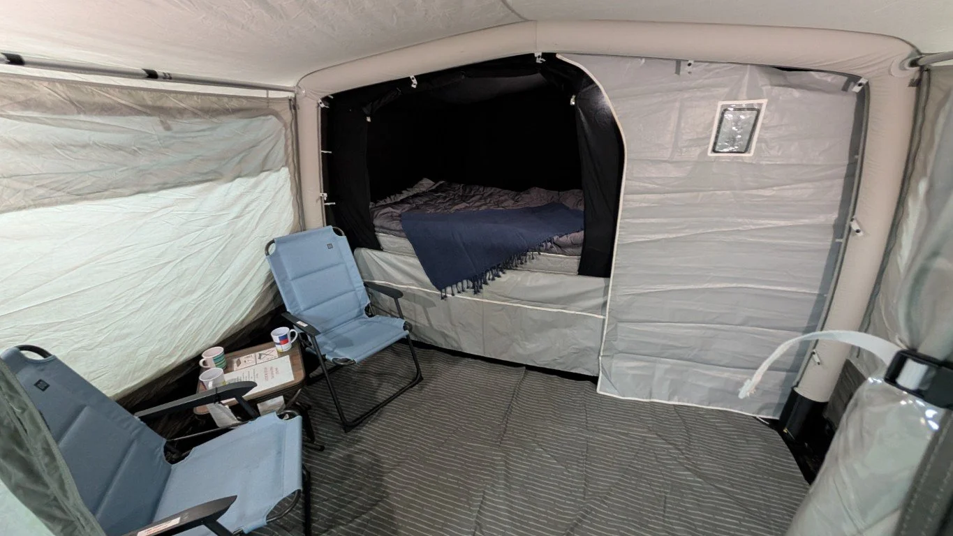 Interior of a camping tent with two chairs, a table with cups and a pamphlet, and a sleeping area with a bed and dark curtains.