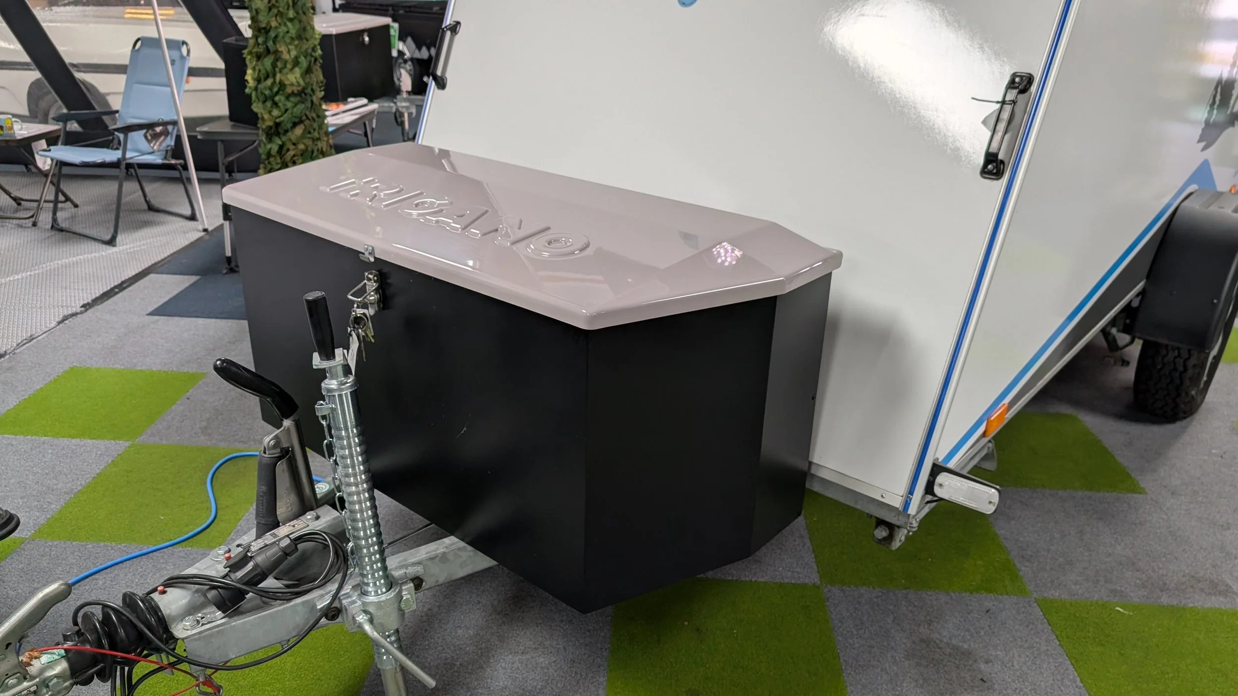 A black and gray toolbox with a shiny lid featuring the raised word 'VIKING' on it, attached to a trailer hitch. The trailer is white with blue accents and parked indoors on a patterned carpet with green squares.