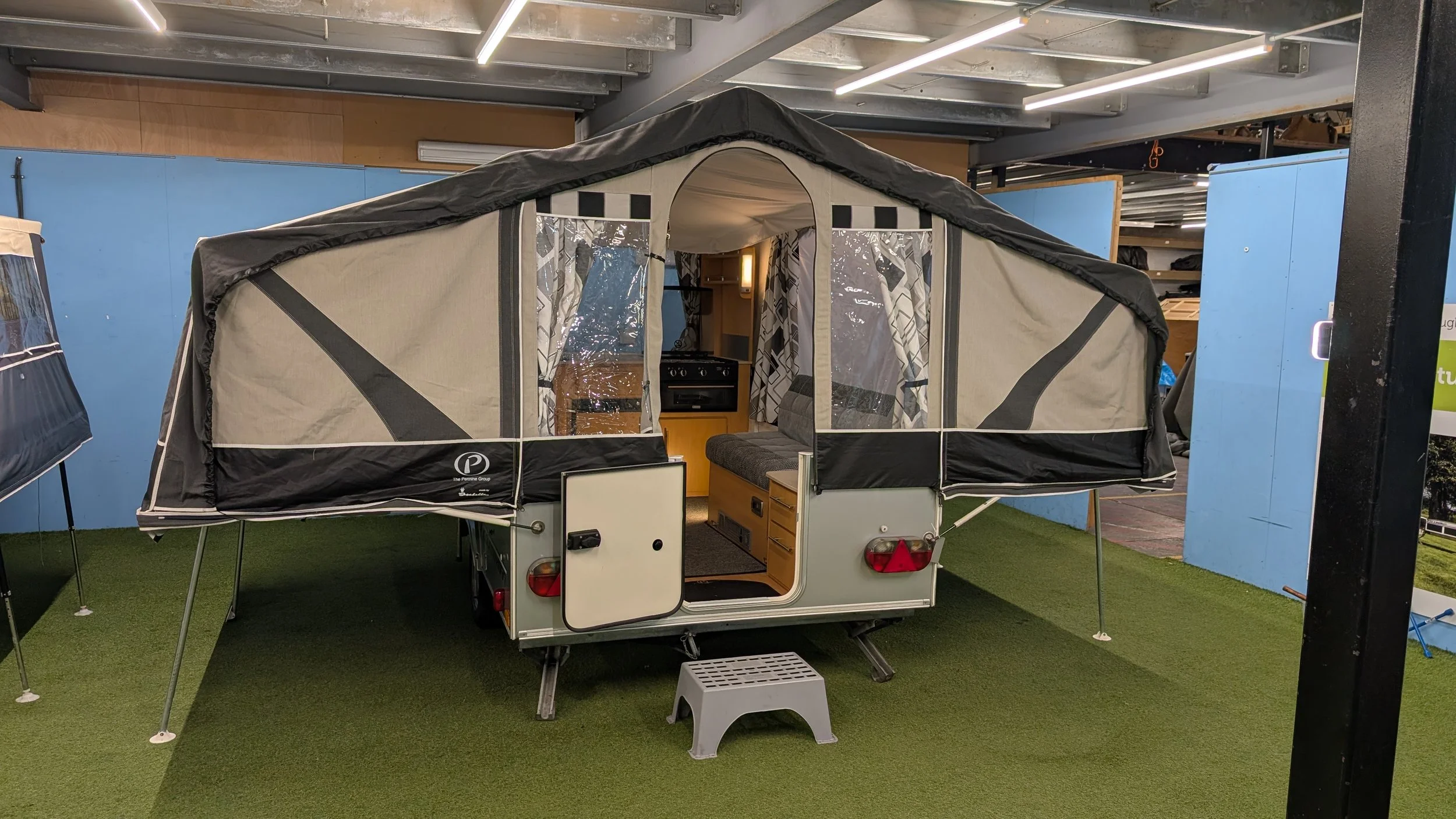 A small camper trailer with a pop-up tent extension set up indoors, with a kitchenette inside and seating area visible.