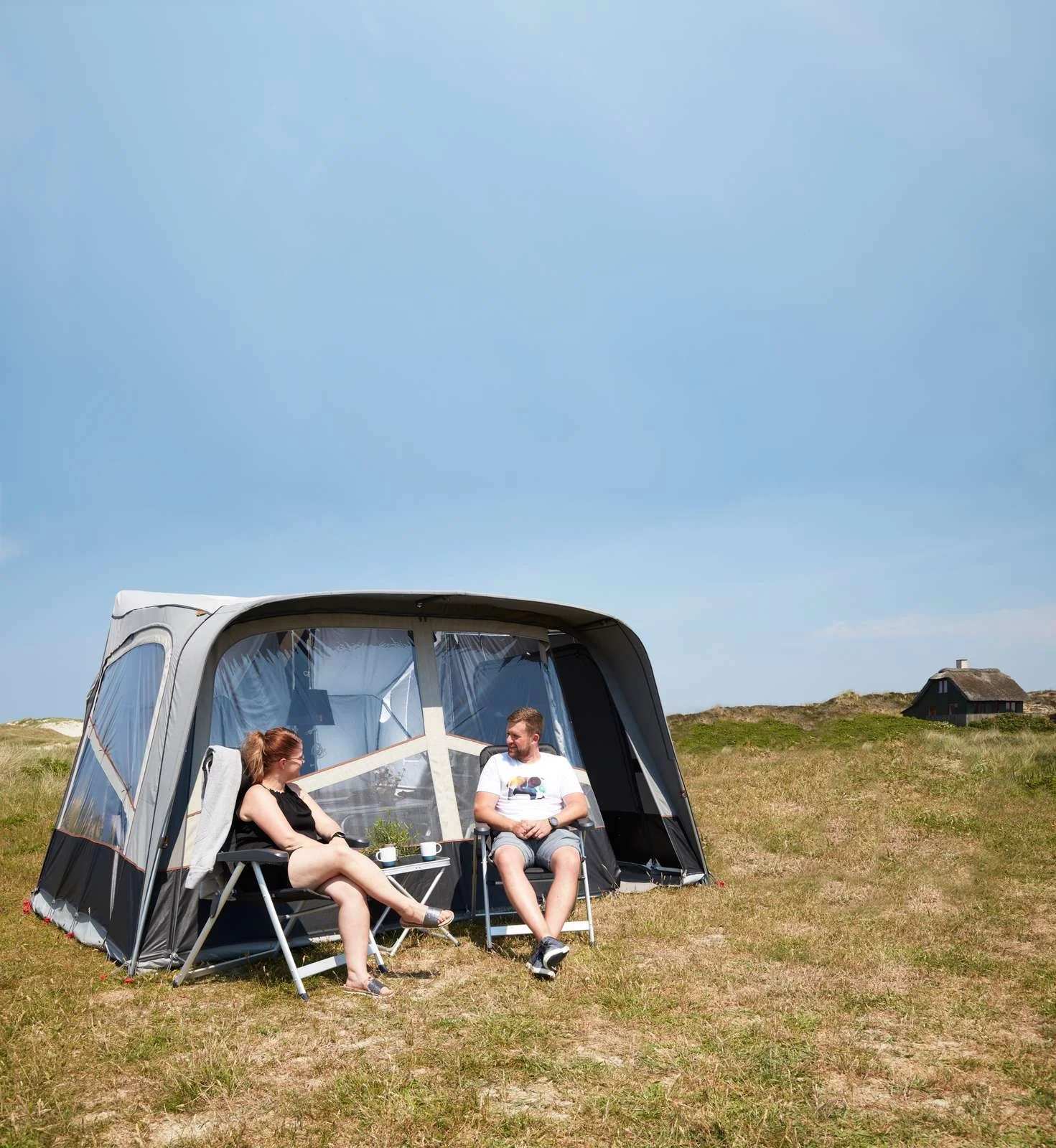 A couple sitting outside a Camp-let tent on a grassy field under a clear blue sky, with a small house in the background.