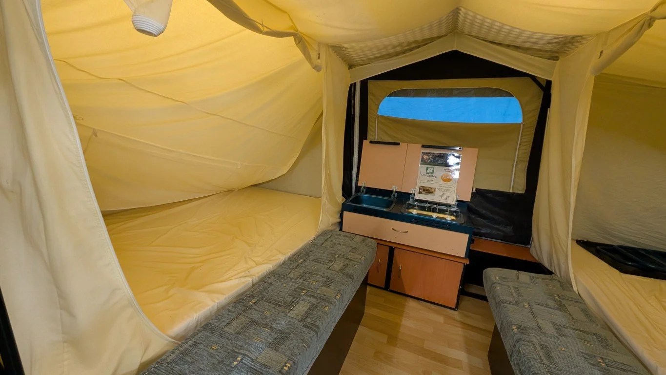Inside a small camping tent with a bunk bed on one side, a bench, and a small kitchenette with a sink and stove.