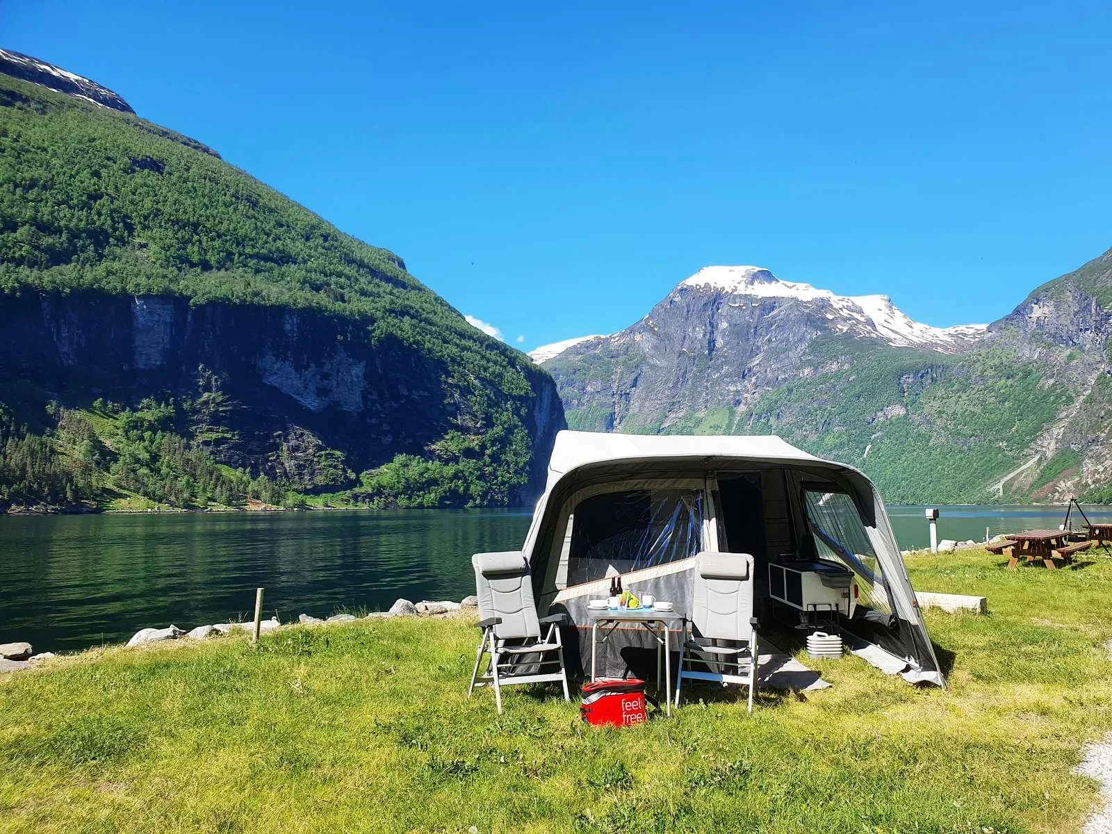 Camp-let tent set up near a lake with mountains and green hills in the background, blue sky, picnic table, chairs, and camping supplies.