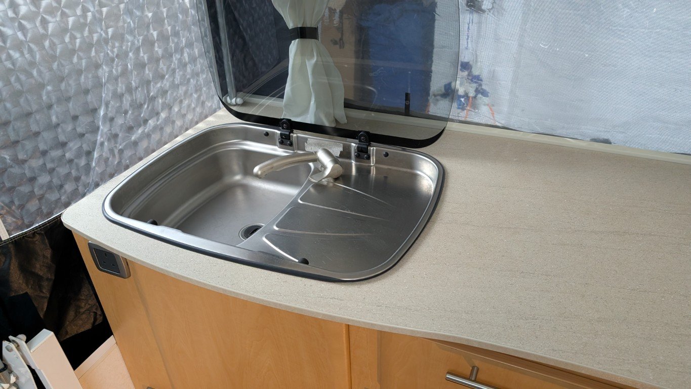 A small kitchen sink with a stainless steel faucet and drain, set in a beige countertop inside a compact living space or camper. There is a window with a retractable shade or curtain above the sink, and a power outlet on the side of the cabinet.