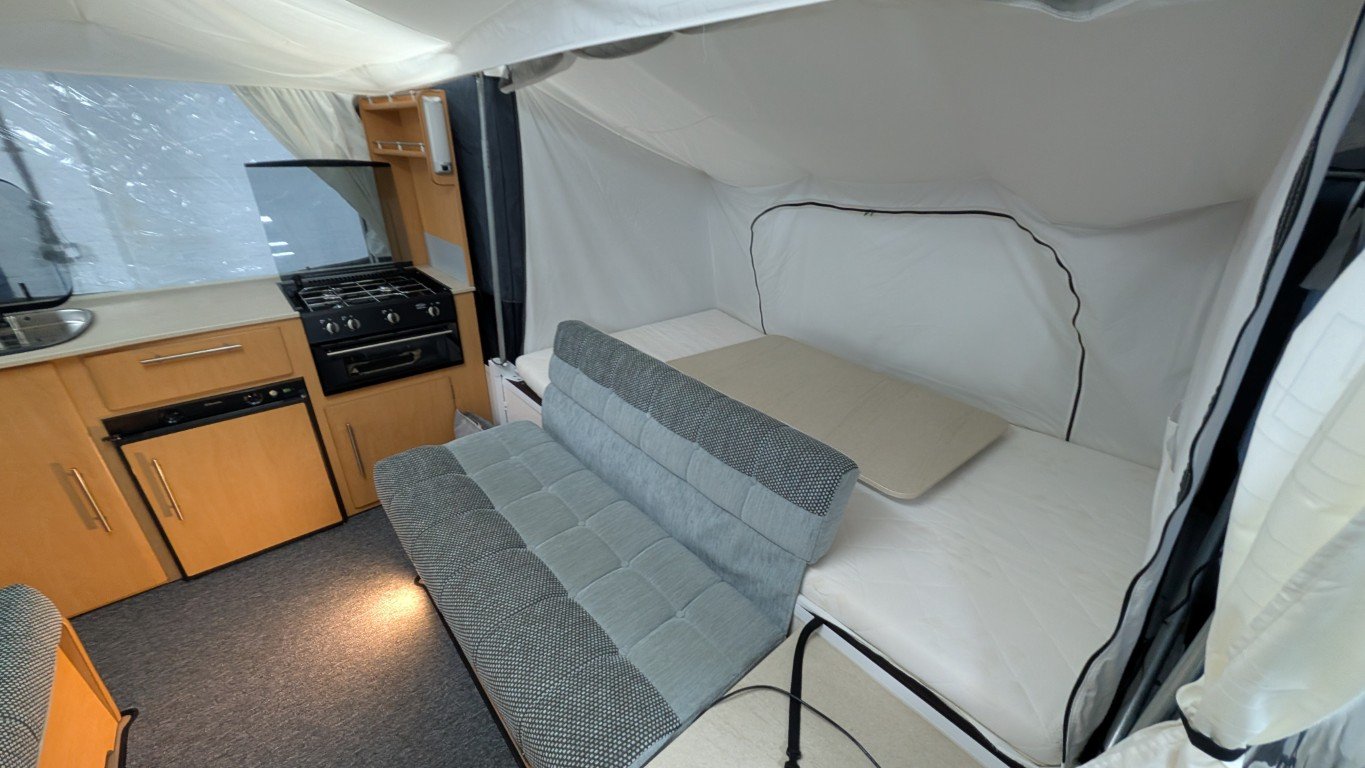 Interior of a small camper or RV with a bed, a fold-out sofa, a small kitchen with a stove, sink, and refrigerator, and a window with curtains.