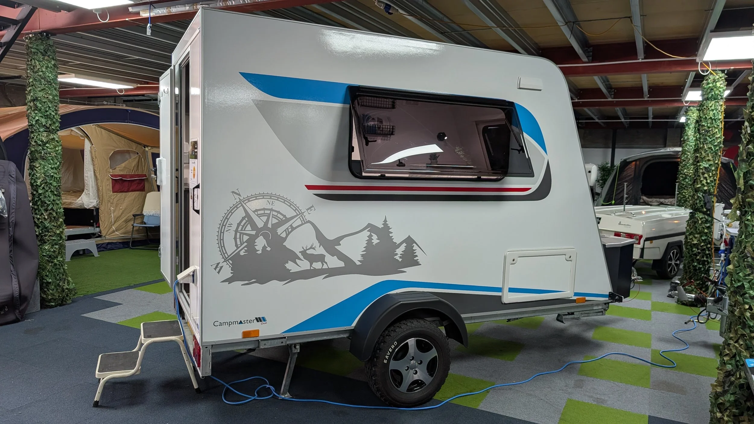 A white camper trailer at an indoor showroom with a mountain and trees graphic, blue and gray accents, and a window. Surrounding it are tents, camping gear, and other trailers, with green and gray flooring and ceiling lights overhead.