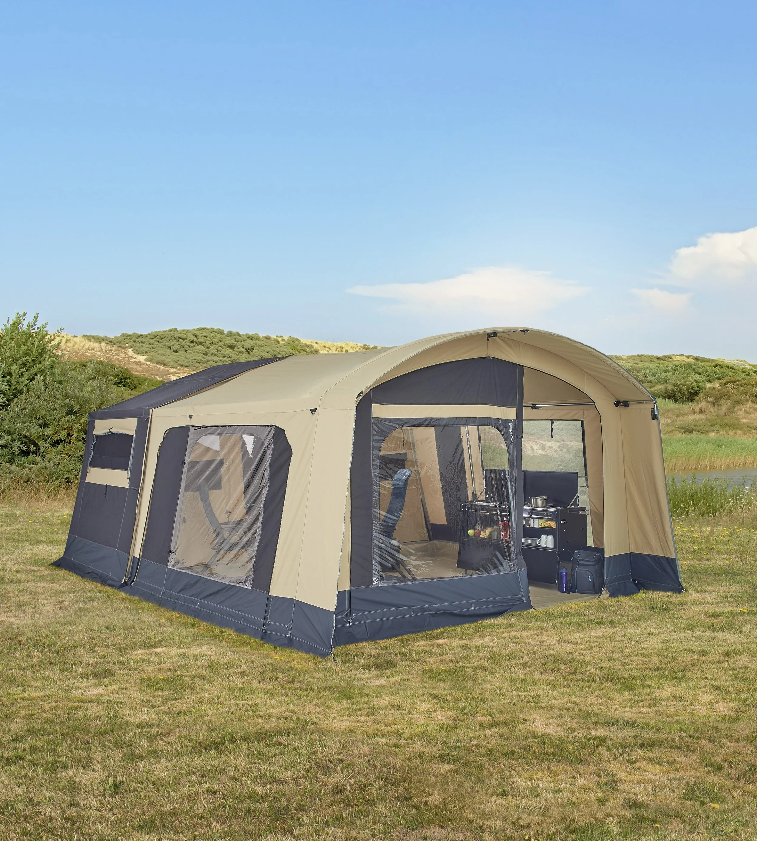 A beige and black Trigano Galleon tent set up on grass with outdoor scenery, blue sky, and some clouds in the background, and a small pond nearby.