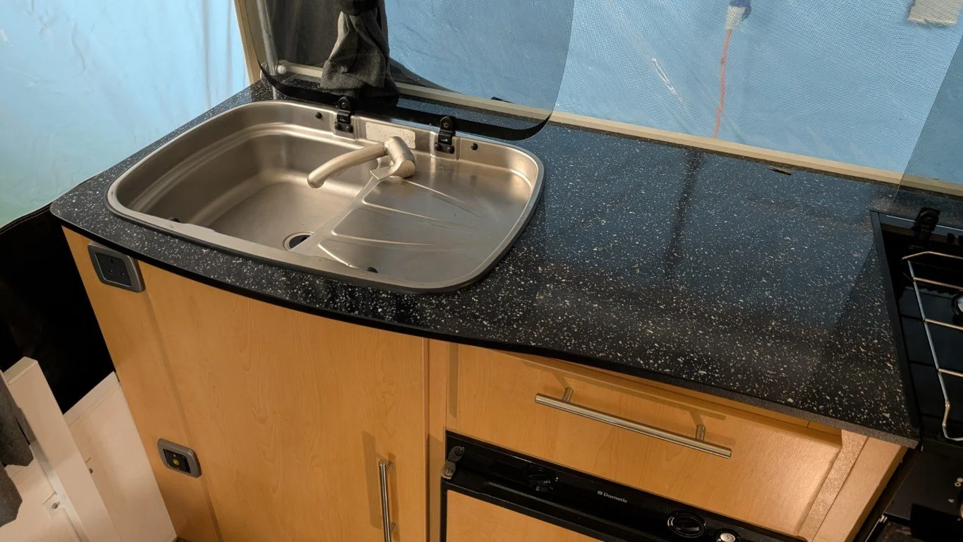 A kitchen countertop with a stainless steel sink, a faucet, and a clear plastic shield behind the sink. The countertop is black with white speckles, and there are wooden cabinets below.