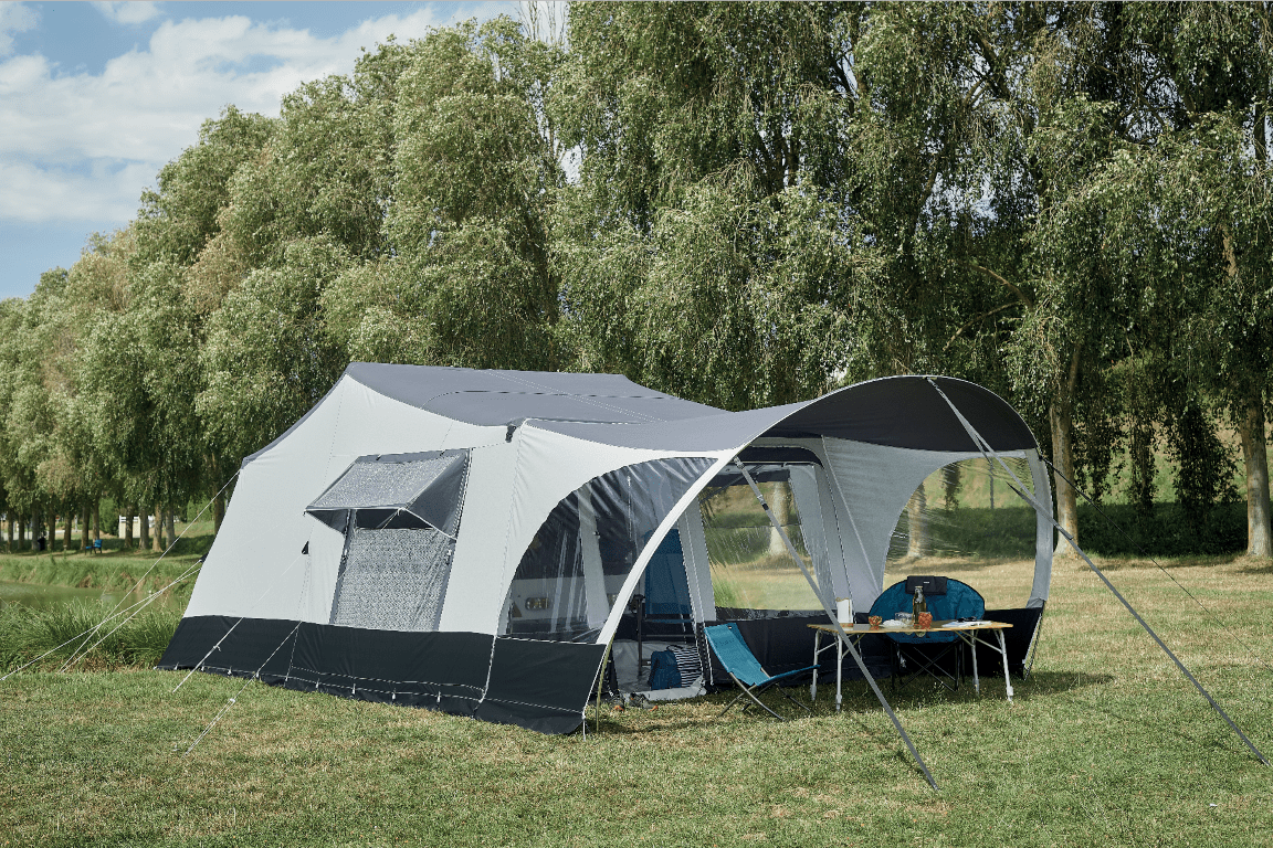 A large outdoor Jamet camping tent set up on a grassy field with trees in the background, with a table and chairs underneath the tent.